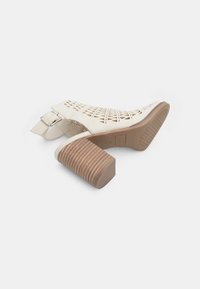 Beige sandali z odprtimi prsti z izrezljanim dizajnom in masivnim lesenim peto. Imajo srebrno kovinsko sponko na zapestju.