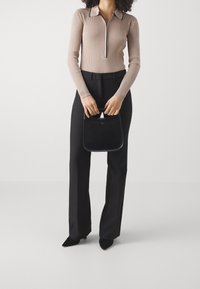 Beige ribbet langærmet top med lynlås ved halsen og sort kant, parret med skræddersyede sorte bukser og holder en lille sort håndtaske.
