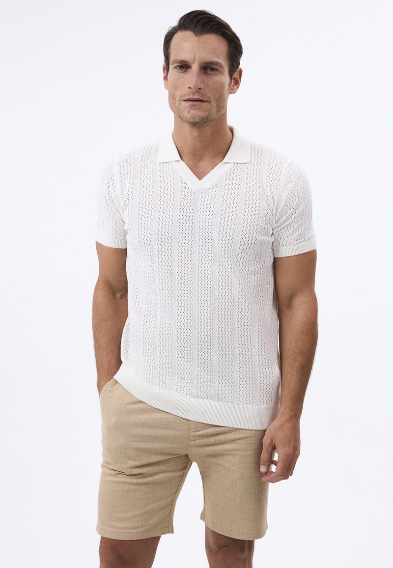 Polo de punto blanco de manga corta con cuello en V, que presenta un patrón texturizado. Combinado con pantalones cortos beiges, enfatizando un aspecto ligero y transpirable.