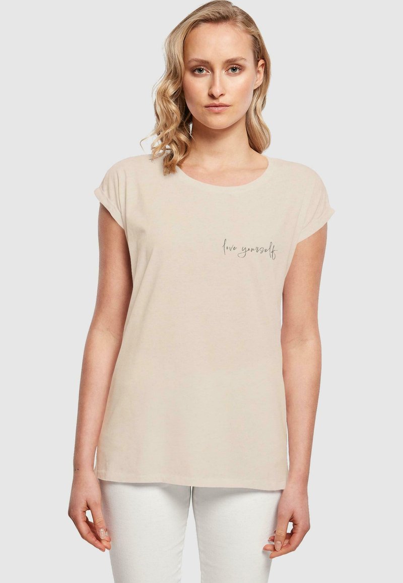 Kortermet beige t-skjorte laget av mykt stoff, med rund hals og en subtil "elsk deg selv" grafikk foran.