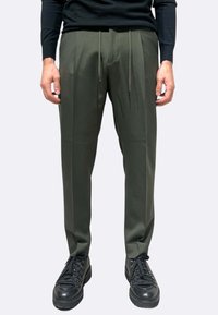 Pantaloni sartoriali verde oliva con vita a coulisse, caratterizzati da gambe dritte e due pieghe frontali, abbinati a scarpe da ginnastica nere con lacci.