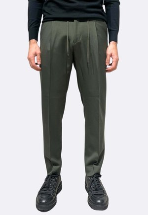 Pantaloni sartoriali verde oliva con vita a coulisse, caratterizzati da gambe dritte e due pieghe frontali, abbinati a scarpe da ginnastica nere con lacci.