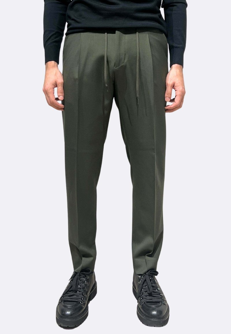 Pantaloni sartoriali verde oliva con vita a coulisse, caratterizzati da gambe dritte e due pieghe frontali, abbinati a scarpe da ginnastica nere con lacci.
