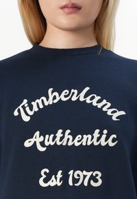 Sötétkék pulóver, fehér hímzett szöveggel, melynek felirata "Timberland Authentic Est 1973", íves kialakításban. Puha szövet textúra.