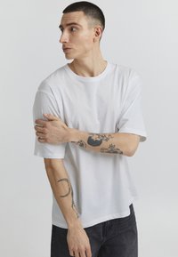 White cotton t-shirt with short sleeves, relaxed fit, crew neck, and visible stitching details. Model posing with arms crossed, showcasing tattoos.