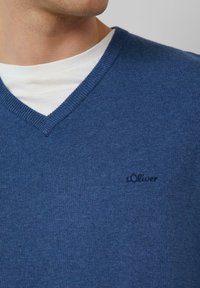 Niebieski sweter w serek wykonany z teksturowanej dzianiny. Posiada małe, haftowane logo na piersi i jest noszony na białej koszuli.