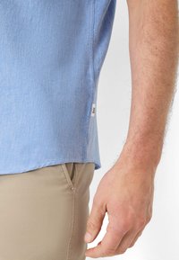 Lichtblauw shirt met korte mouwen van een gestructureerde stof, voorzien van een zijsplit en een klein merkembleem, gecombineerd met kaki broek.