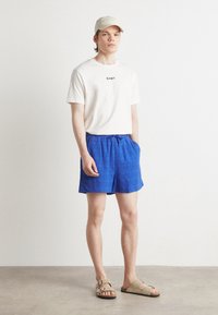 Camiseta blanca con el logotipo de "GANT", pantalones cortos azules con estampado y sandalias beige. El sujeto lleva una gorra clara y está de pie sobre un suelo neutro.