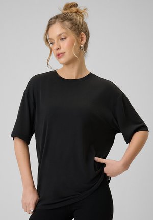 Kobieta z blond włosami związanymi w kok, nosząca srebrne kolczyki koła i luźną czarną koszulkę z krótkim rękawem, pozująca z jedną ręką na biodrze.