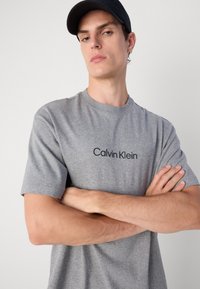 Grå bomull T-skjorte med korte ermer, med en svart "Calvin Klein"-logo på forsiden. Modellen krysser armene og har på seg en svart caps.