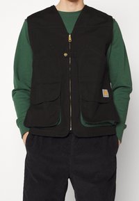 Schwarze Canvas-Weste mit Reißverschluss und goldener Hardware. Verfügt über zwei große Fronttaschen. Wird über einem grünen Langarmshirt getragen.