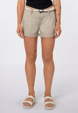 Person trägt beige Shorts mit umgeschlagenem Saum und weißem geflochtenem Gürtel, schwarzes Top, beige Sandalen und sichtbare Tattoos an Armen und Beinen.