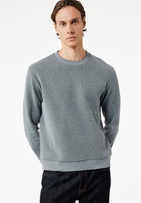 Sudadera gris con canalé, cuello redondo, mangas largas y un acabado texturizado, combinada con jeans oscuros.