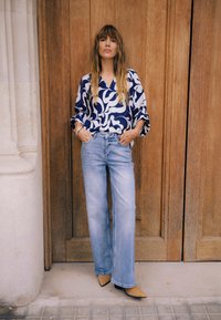 Lahtise lõikega tumesinise ja valge mustriga pluus, helerohelised laia säärega teksad ja beežid teravnurgaga jalanõud. Taustal on puidust uksed.