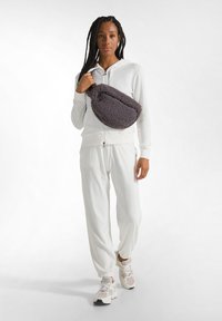 Completo atletico bianco con giacca con cappuccio e zip e pantaloni affusolati. Il modello porta una borsa morbida grigia, completata da sneakers crema.