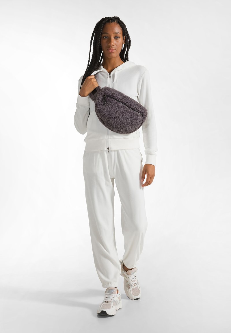 Completo atletico bianco con giacca con cappuccio e zip e pantaloni affusolati. Il modello porta una borsa morbida grigia, completata da sneakers crema.