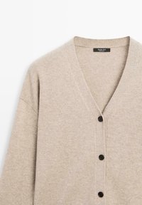 Beige stickad kardi med V-ringning, långa ärmar och fem svarta knappar. Mjuk textur med subtila sömdetaljer.