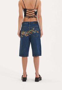 Denim bermuda shorts with a dark wash, featuring intricate multicolored dragon embroidery on back pockets and rolled hems.