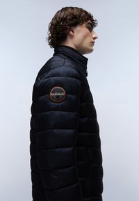 Sort pufferjakke med høj krave, quiltet design og en sidelomme med lynlås. Har et rundt mærke på venstre arm.