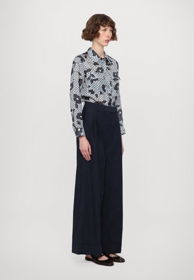 Langärmliges Lichtblaues Hemd mit einem geometrischen und floralen Muster. Hochtaillierte weite Hosen aus dunkelblauem Denim. Braune flache Schuhe.