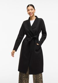 Cappotto lungo nero con colletto a scialle, cintura in vita e tasche laterali. Indossato sopra pantaloni ampi con stampa animale. Texture liscia.