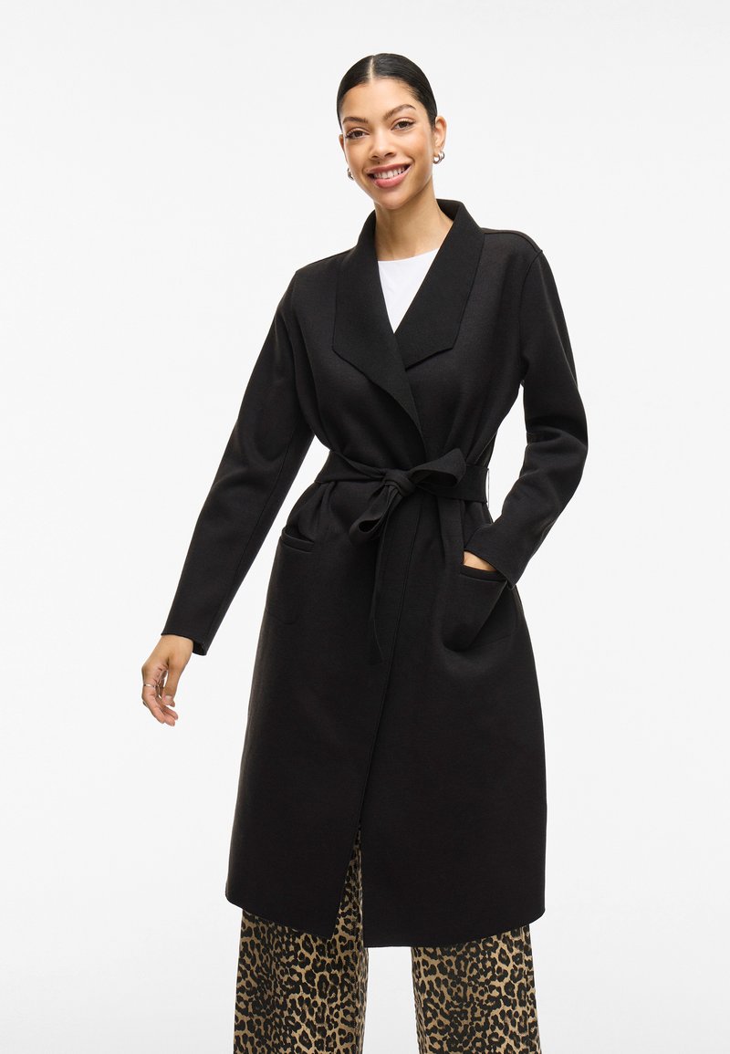 Cappotto lungo nero con colletto a scialle, cintura in vita e tasche laterali. Indossato sopra pantaloni ampi con stampa animale. Texture liscia.