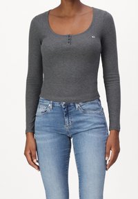 Graues langärmliges Rippoberteil mit weitem Rundhalsausschnitt, einer dreiknöpfigen Knopfleiste und figurbetontem Schnitt, kombiniert mit hellblauen Jeans.