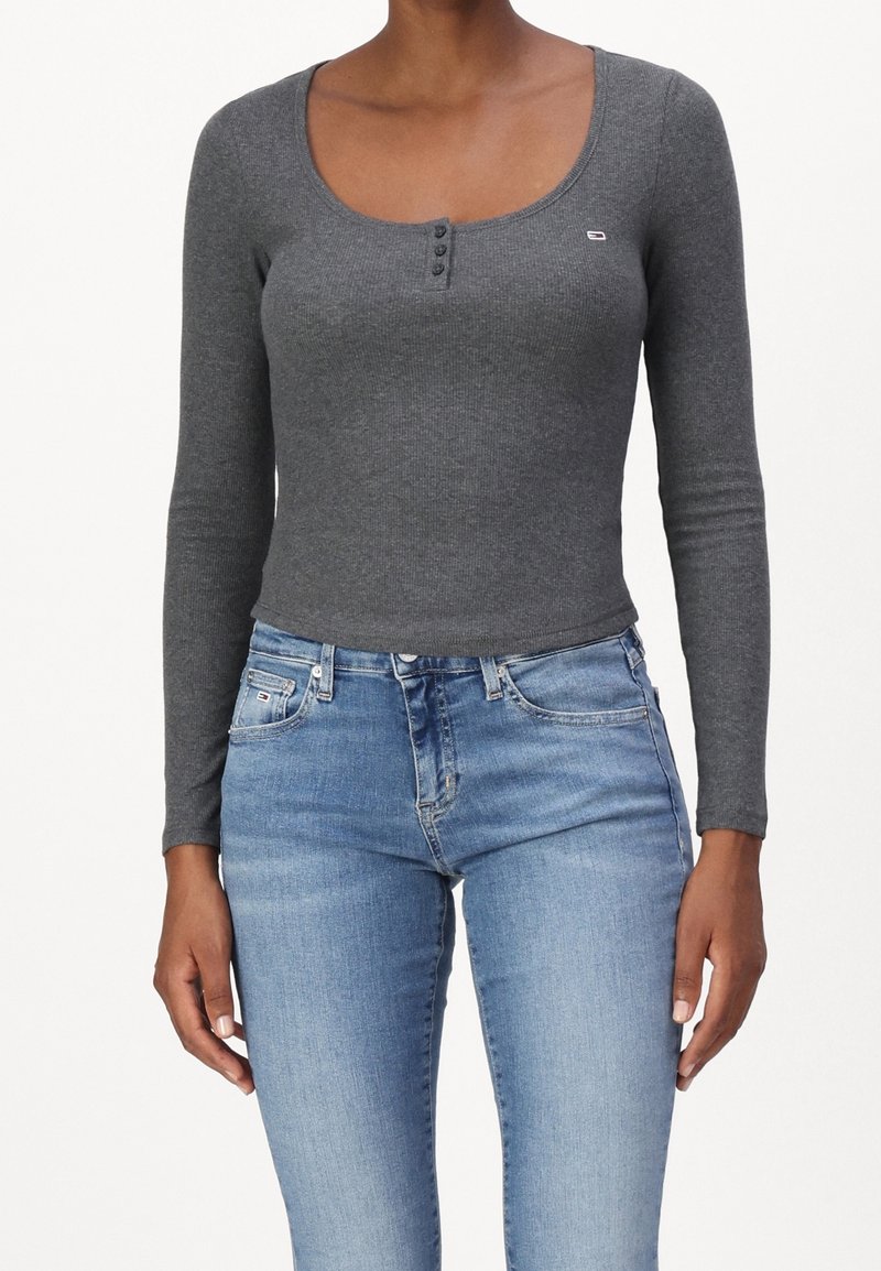 Graues langärmliges Rippoberteil mit weitem Rundhalsausschnitt, einer dreiknöpfigen Knopfleiste und figurbetontem Schnitt, kombiniert mit hellblauen Jeans.