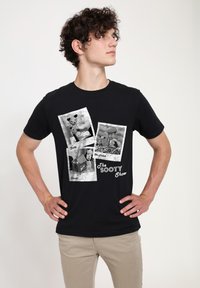 Svart bomull t-shirt med tre tryckta bilder i vintage-stil som föreställer karaktärer från "The Sooty Show", med kontrasterande vit text.