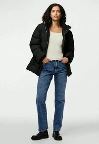 Črna puhovka s kapuco, sprednjim zadrgo in gumbi, nosena čez bež majico in modre kavbojke. Prilagojeno in teksturirano blago, v kombinaciji s črnimi čevlji.