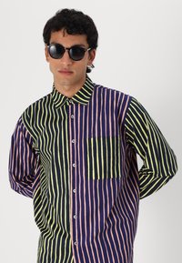 Camisa de botón a rayas con rayas verticales en azul marino, rosa y verde; incluye un bolsillo en el lado izquierdo; textura de tela suave.