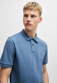 Jasnoniebieska koszulka polo z teksturowanej tkaniny, z kołnierzykiem, trzema guzikami z przodu oraz małą naszywką z logo na piersi.