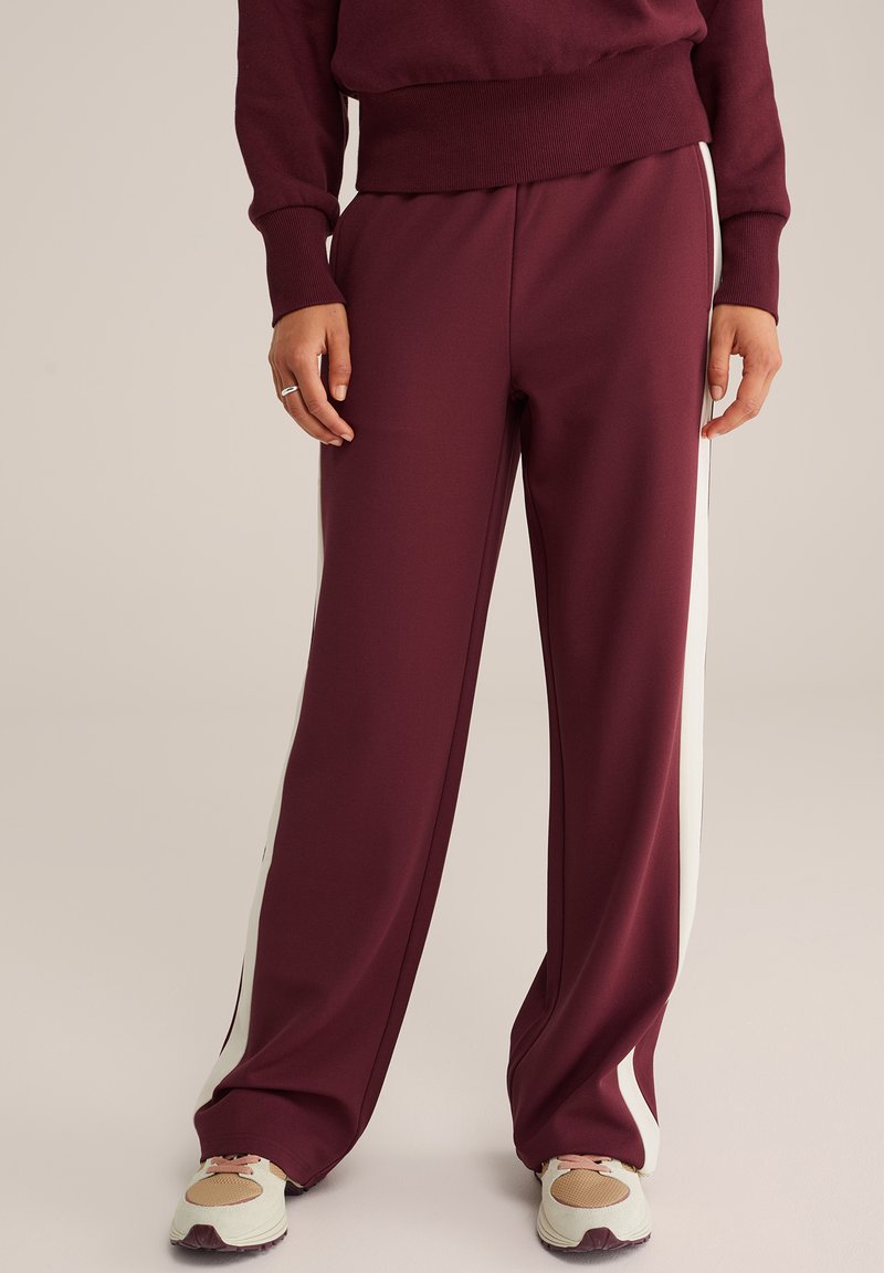 Bordó tréningnadrág puha anyagból, fehér oldalsávokkal, laza szabással és elasztikus derékkal. Sportcipővel párosítva.