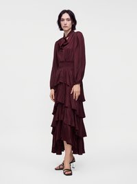 Femme portant une longue robe burgundy à volants avec un col haut à nœud et des manches longues, associée à des sandales à talons noires à brides.