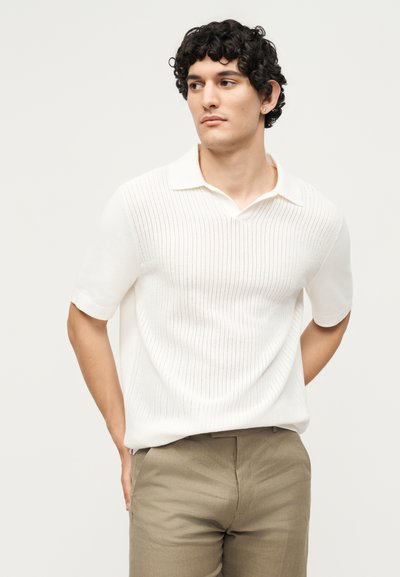 Άνδρας με σγουρά σκούρα μαλλιά που φοράει λευκό πλεκτό πόλο με κοντά μανίκια και μπεζ παντελόνι, κοιτάζοντας αριστερά με τα χέρια πίσω από την πλάτη.