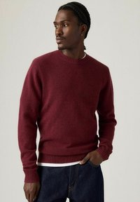 Bordeaux ronde hals trui van zachte stof, ontworpen met geribbelde mouwen en zoom, gedragen over een wit shirt, gecombineerd met donkere jeans.