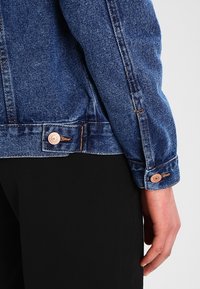 Primer plano del brazo de una persona que lleva una chaqueta de denim azul con botones de cobre, con la mano descansando a un lado contra un fondo de pantalones negros.