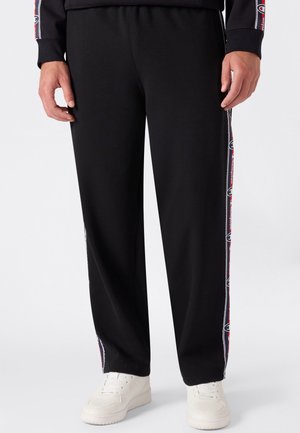Persona che indossa pantaloni neri con strisce rosse e bianche con logo sui lati e scarpe da ginnastica bianche, in piedi con le braccia rilassate lungo i fianchi.