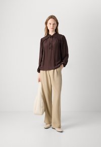 Brun plisseret bluse med sløjfe, beige vidde bukser og lyse sko, kompletteret af en beige tote taske. Glat stofstruktur.