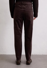 Bruine corduroy broek met een recht model, achterzakken en een gestructureerde afwerking. Draagt met donkere schoenen tegen een neutrale achtergrond.