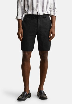 Mann trägt schwarze knielange Shorts, schwarze Slipper und ein weißes Hemd mit vertikalen schwarzen Streifen.