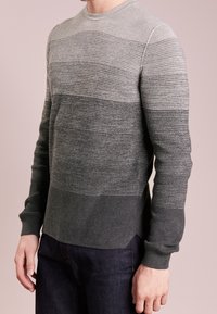 Homme portant un pull en maille gris dégradé avec des poignets côtelés et un jean noir sur un fond rose pâle uni.