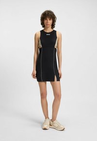 Schwarzes ärmelloses Kleid mit ausgeschnittenen Seiten und beigen Akzenten. Verfügt über weiße Paspelierung und einen seitlichen Schlitz. Kombiniert mit beigen Sneakers.