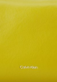 Dzeltena ādas virsma ar strukturētu apdari, ar "Calvin Klein" izcirstu baltā krāsā apakšējā centrā.
