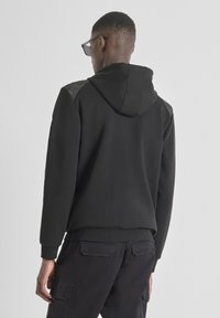 Sudadera negra con un panel texturizado gris en los hombros, que presenta una capucha con cordón, puños acanalados y un corte relajado.