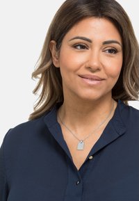 Silberne Halskette mit einem rechteckigen Anhänger, der über einem dunkelblauen Hemd mit Kragen getragen wird. Der Anhänger weist geprägte Details auf.