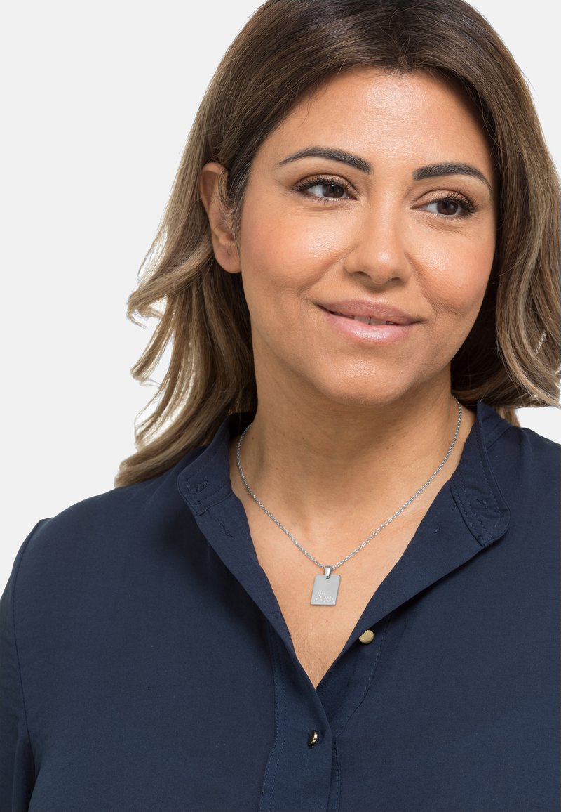 Silberne Halskette mit einem rechteckigen Anhänger, der über einem dunkelblauen Hemd mit Kragen getragen wird. Der Anhänger weist geprägte Details auf.