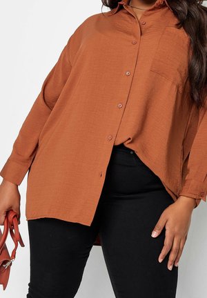 Femme portant une chemise ample couleur rouille boutonnée et un pantalon noir, tenant un petit sac à main assorti couleur rouille à ses côtés.