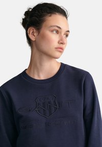 Felpa blu navy con collo rotondo, caratterizzata da un logo GANT in rilievo e la scritta "AMERICAN SPORTSWEAR". Tessuto morbido e dalla texture particolare.
