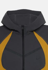 Tamno siva dukserica s prednjim zipom, senf žuti bočni paneli i mali crni Nike logo na grudima.